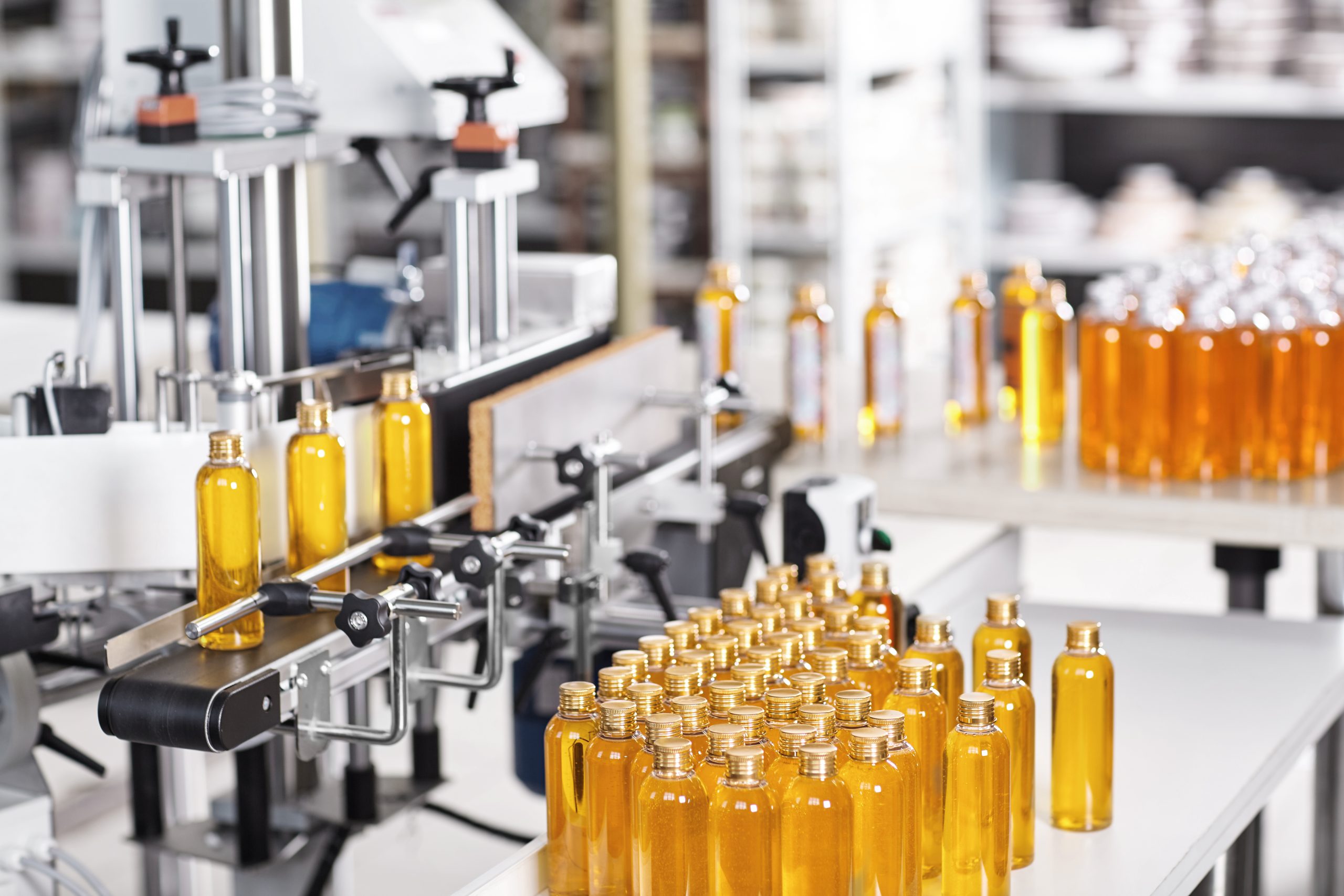 Bottles filled with yellow thick substance standing on desk and conveyor line. Production line of cosmetics products on big factory.Automated process of creation shampoo on factory.